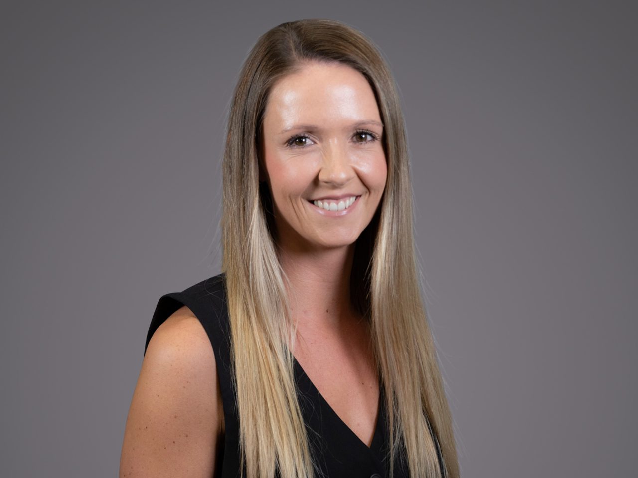 A person with long blonde hair, wearing a sleeveless black top, smiling at the camera against a grey studio backdrop.