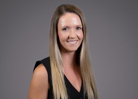 A person with long blonde hair, wearing a sleeveless black top, smiling at the camera against a grey studio backdrop.