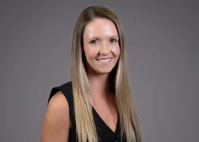A person with long blonde hair, wearing a sleeveless black top, smiling at the camera against a grey studio backdrop.