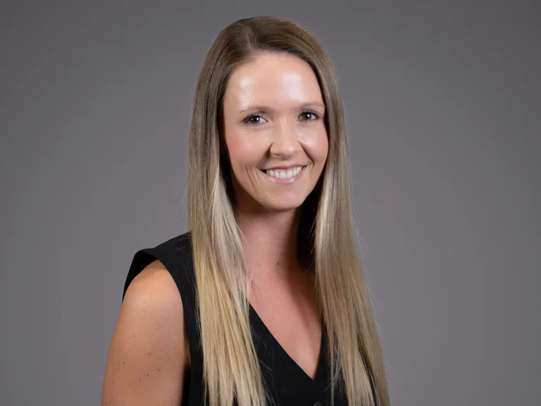 Portrait of a person with long blonde hair, wearing a black sleeveless top, smiling against a neutral gray studio background.