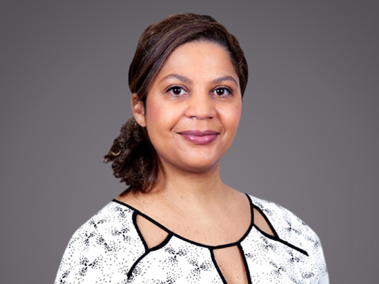 Portrait of a person with dark hair pulled back, wearing a white patterned blouse with black trim, smiling softly against a gray background.