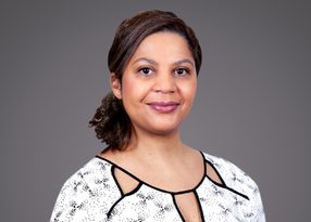 Portrait of a person with dark hair pulled back, wearing a white patterned blouse with black trim, smiling softly against a gray background.
