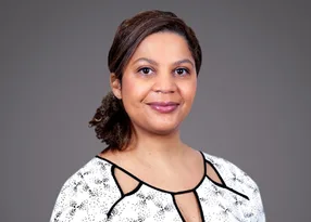 Portrait of a person with dark hair pulled back, wearing a white patterned blouse with black trim, smiling softly against a grey backdrop.