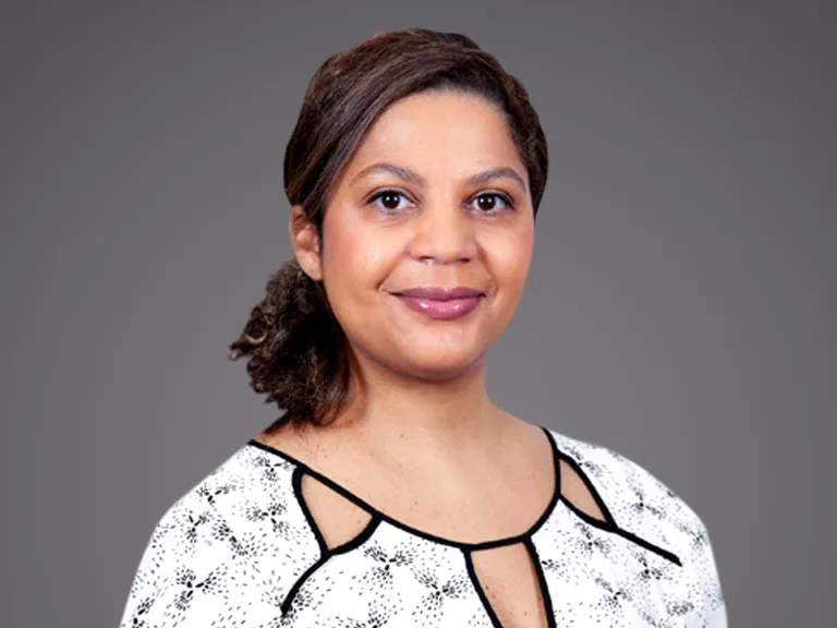 Portrait of a person with dark hair pulled back, wearing a white patterned blouse with black trim, smiling softly against a grey backdrop.