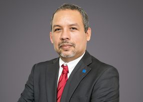 Portrait of a person in a dark suit, white shirt and red tie, with a small blue lapel pin, against a plain grey background.