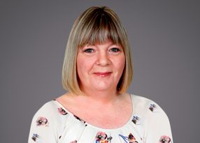 Portrait of a person with shoulder-length blonde hair and bangs, wearing a light floral blouse, smiling against a gray background.