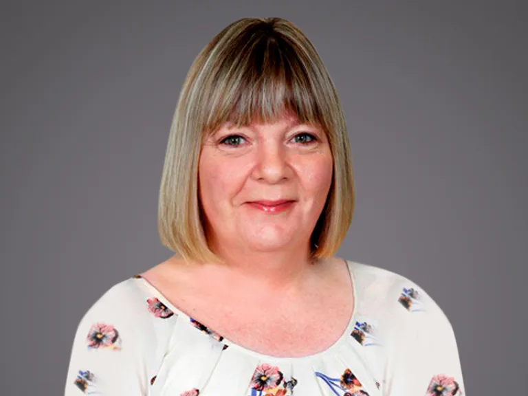 Persona con capelli biondi corti a caschetto e frangia, sorriso leggero, indossa camicia bianca con motivo floreale, sfondo grigio.