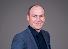 Portrait of a smiling person in a blue suit, dark shirt and tie, standing against a neutral grey backdrop.