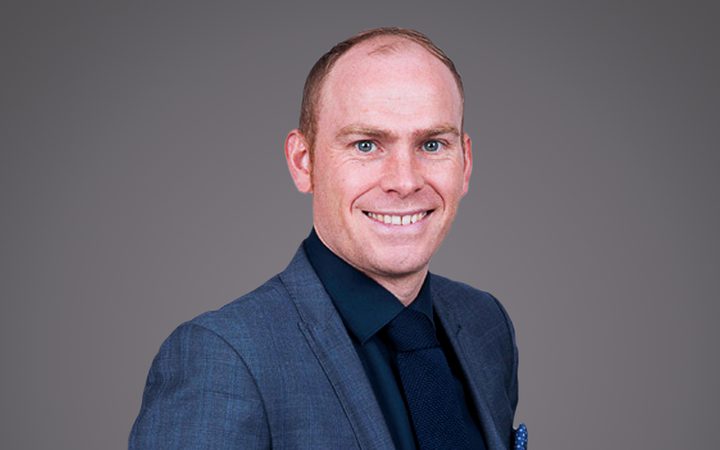 Portrait of a smiling person in a blue suit, dark shirt and tie, standing against a neutral grey backdrop.