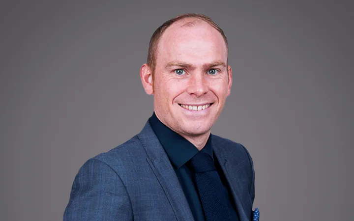 Portrait of a smiling person in a blue suit, dark shirt and tie, standing against a neutral grey backdrop.