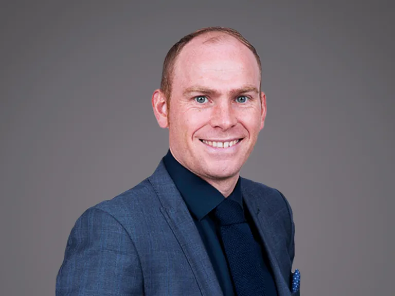 Portrait of a smiling person in a blue suit, dark shirt and tie, standing against a neutral grey backdrop.