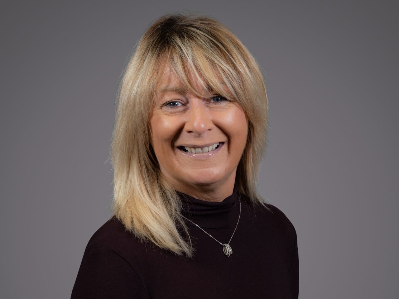 A person with shoulder-length blond hair, smiling, wearing a black turtleneck and a silver pendant necklace, against a plain grey studio backdrop.