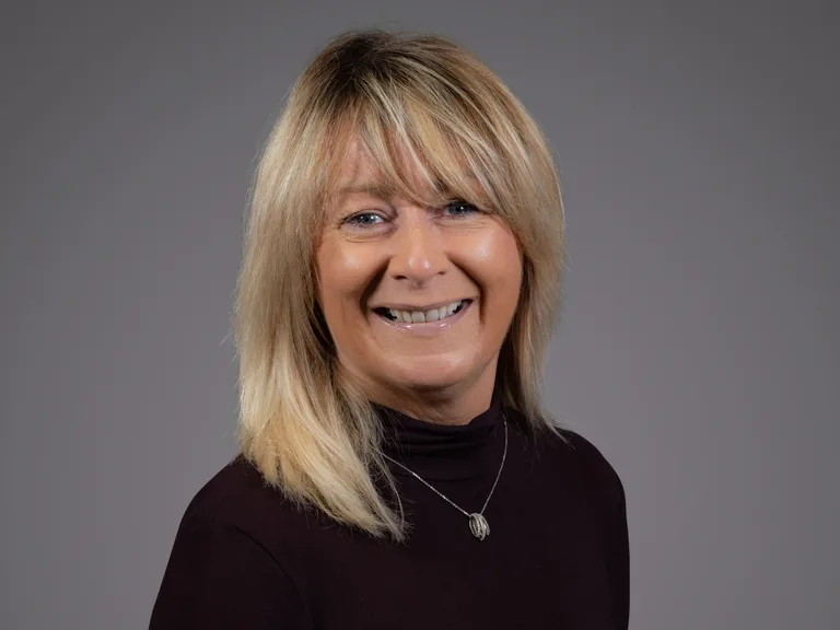 Smiling person with shoulder-length blonde hair, wearing a black turtleneck and a silver pendant necklace, against a gray studio backdrop.