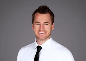 A person with short brown hair smiles at the camera, wearing a white dress shirt and black tie, against a plain gray studio backdrop.