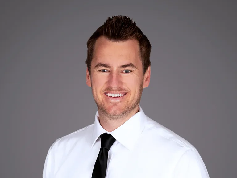 Lächelnde Person in weißem Hemd und schwarzer Krawatte vor grauem Hintergrund.