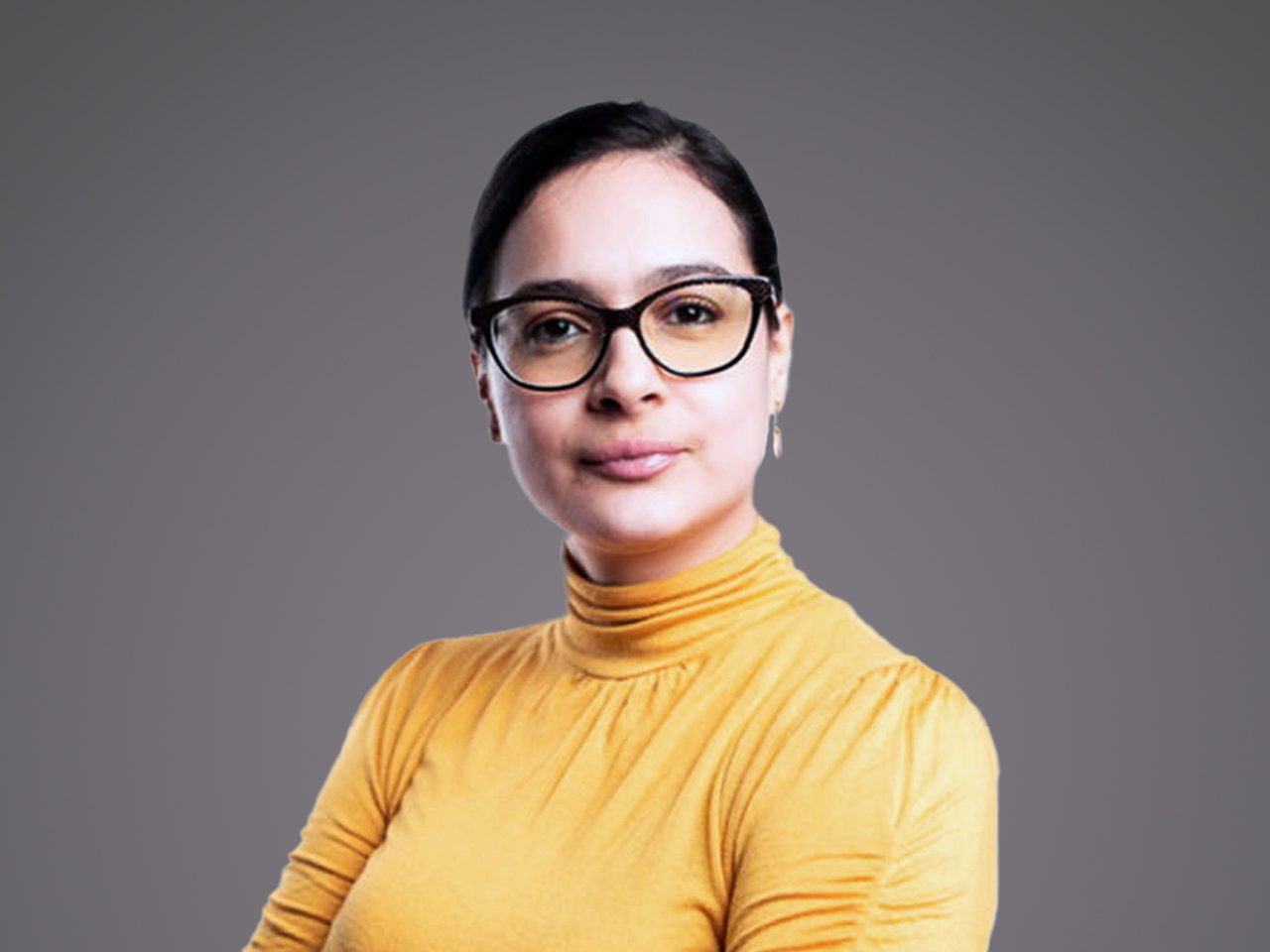 A person with short dark hair and black glasses, wearing a mustard-yellow turtleneck, looks at the camera with a soft smile against a grey backdrop.