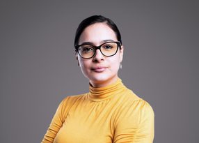 A person with short dark hair and black glasses, wearing a mustard-yellow turtleneck, looks at the camera with a soft smile against a grey backdrop.