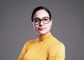 A person with short dark hair, wearing black-framed glasses and a mustard-yellow turtleneck, posed against a gray gradient background.