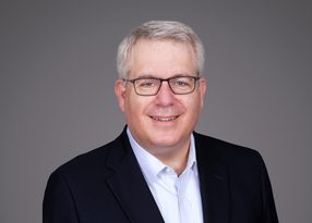 Portrait of a person with short grey hair and glasses, wearing a dark blazer and white shirt, smiling against a plain grey background.