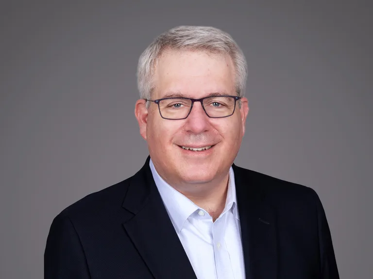 Smiling person with short gray hair and glasses, wearing a dark blazer over a light blue shirt, posed against a plain gray background.