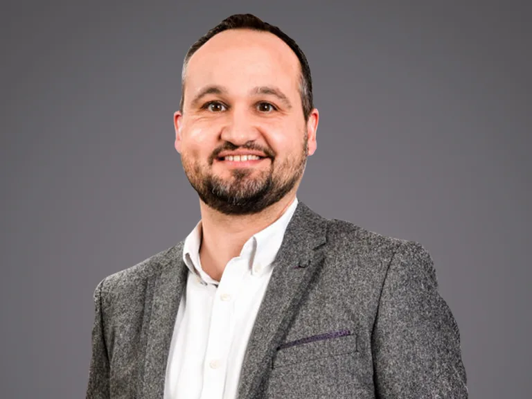 Personne souriante en chemise blanche et veste grise texturée, sur fond gris neutre.