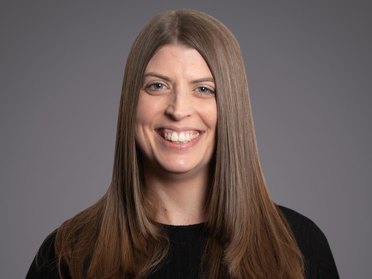 A person with long, straight light brown hair, wearing a black top, smiling against a gray studio backdrop.