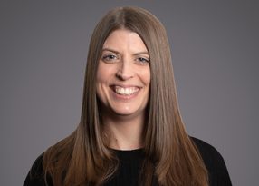 A person with long, straight light brown hair, wearing a black top, smiling against a gray studio backdrop.