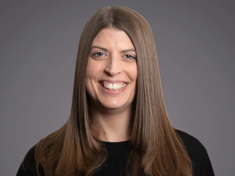 Persona con lunghi capelli castano chiaro, maglia nera, sorriso ampio, sfondo grigio uniforme.