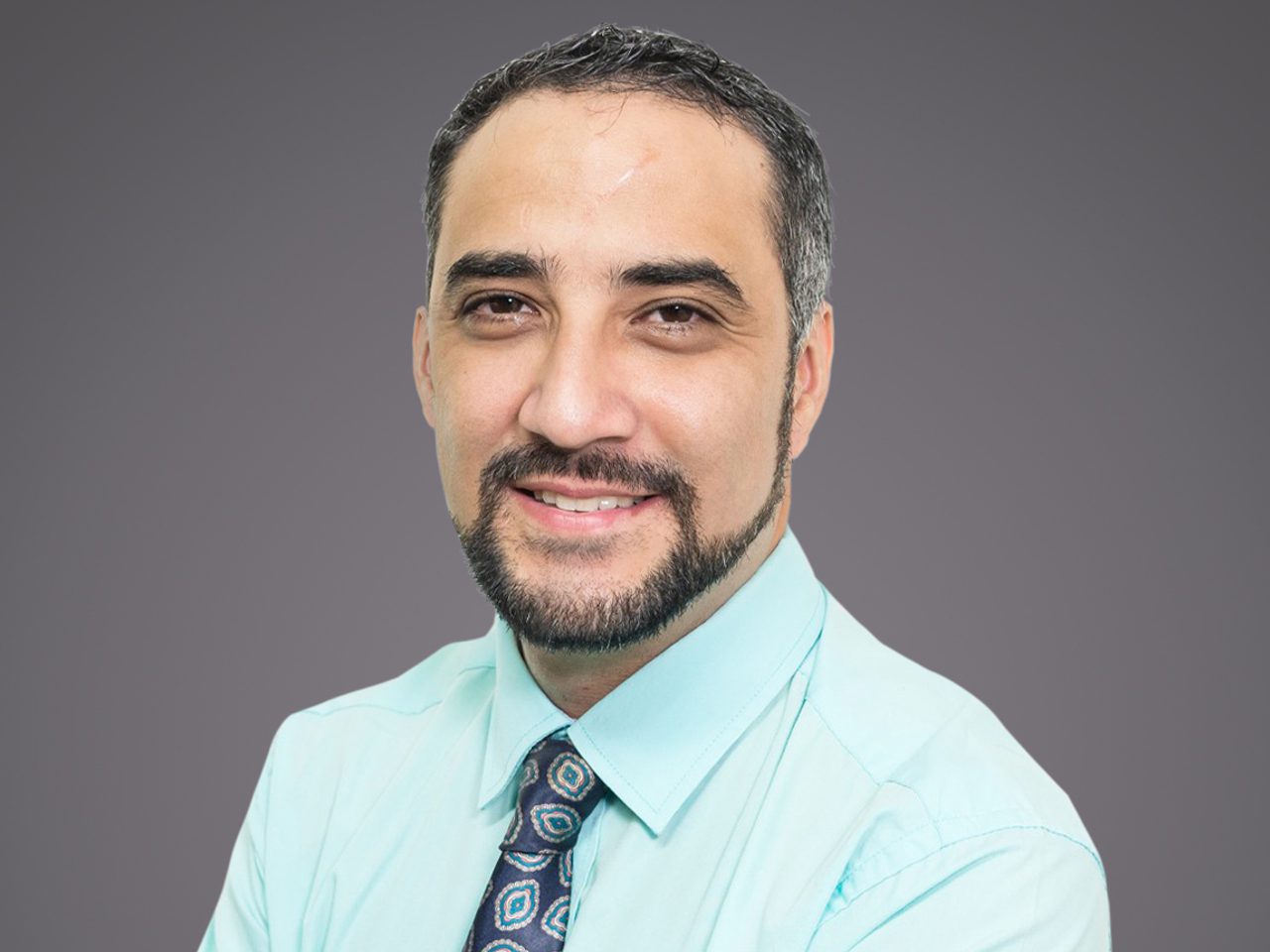 Portrait d'une personne souriante avec barbe et cheveux bruns, portant une chemise bleu clair et une cravate à motifs, fond gris.
