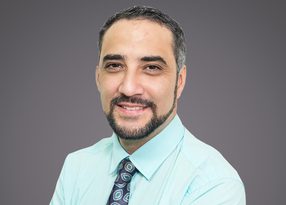 Portrait d'une personne souriante avec barbe et cheveux bruns, portant une chemise bleu clair et une cravate à motifs, fond gris.