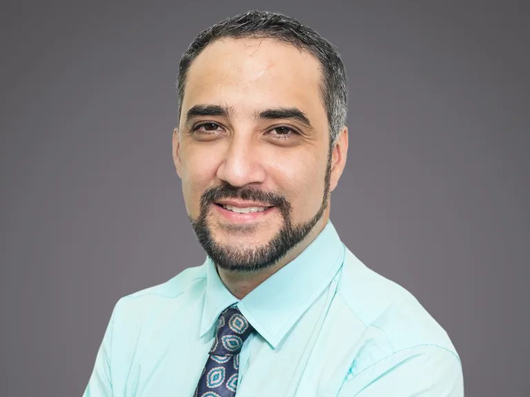 Portrait d'une personne souriante avec barbe et cheveux bruns, portant une chemise bleu clair et une cravate à motifs, fond gris.