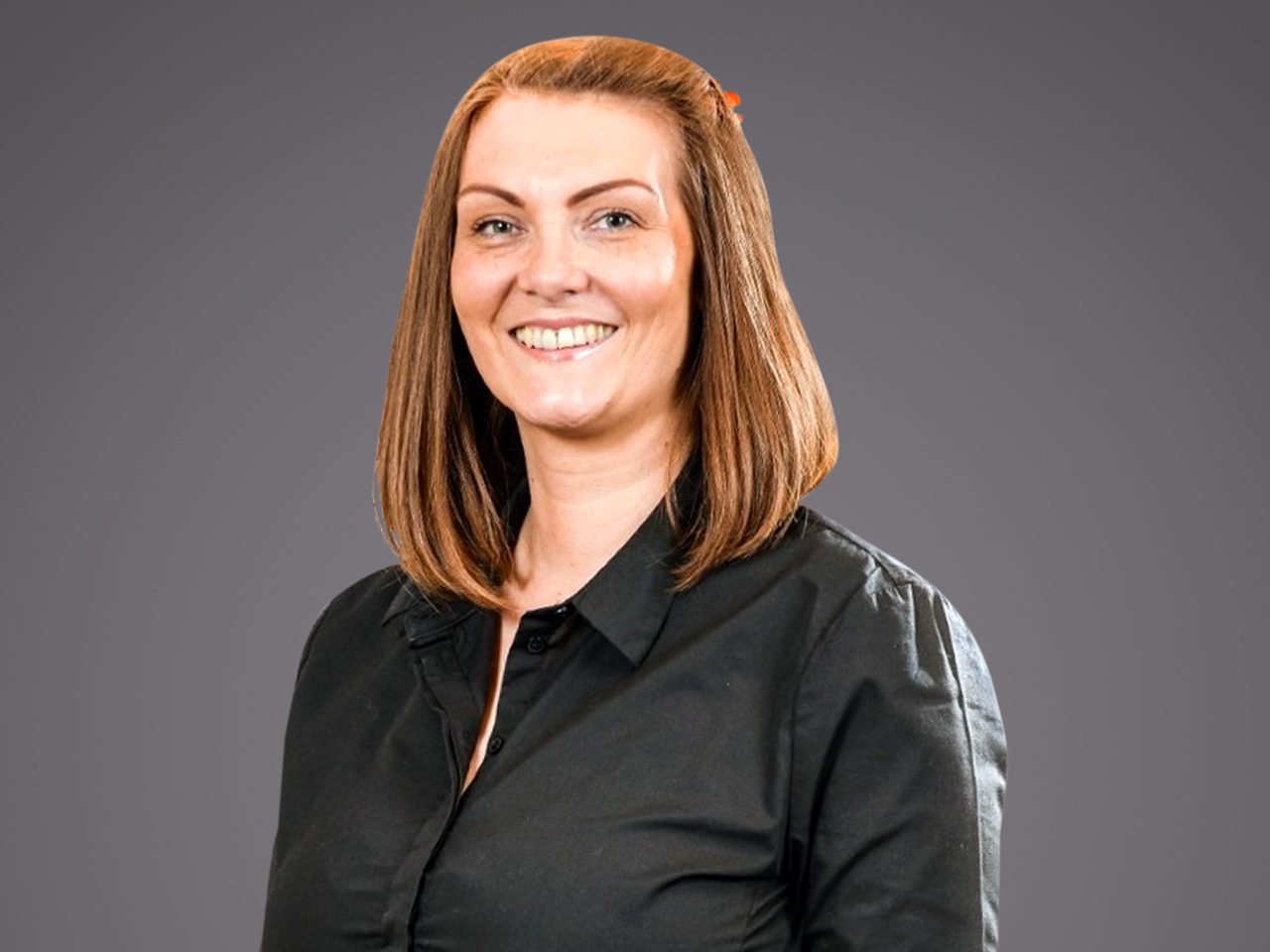 A person with shoulder-length light brown hair, wearing a black blouse, smiling at the camera against a grey studio backdrop.