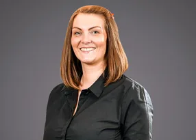 A person with shoulder-length light brown hair, wearing a black blouse, smiling at the camera against a grey studio backdrop.