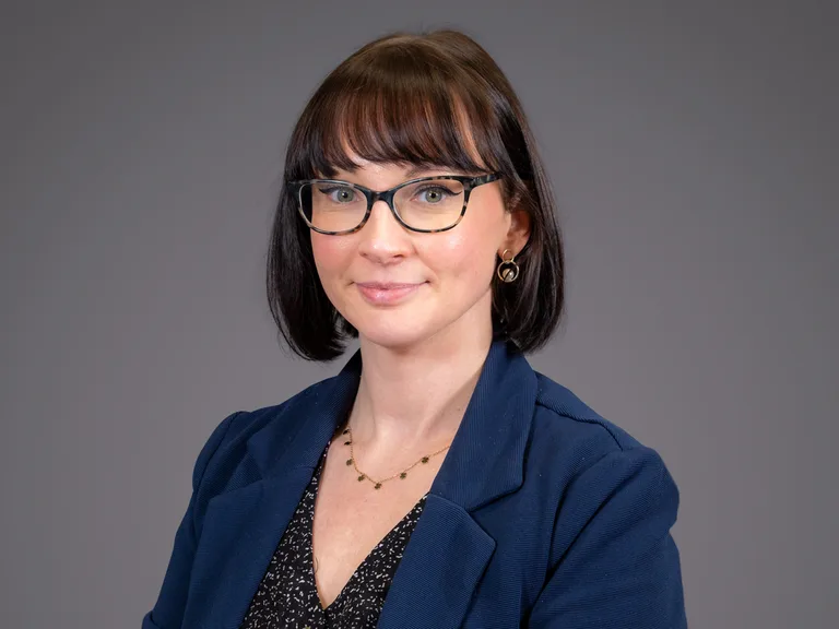 Portrait d'une personne portant lunettes, collier et boucles d'oreilles, veste bleue et chemise sombre, cheveux bruns courts, fond gris.
