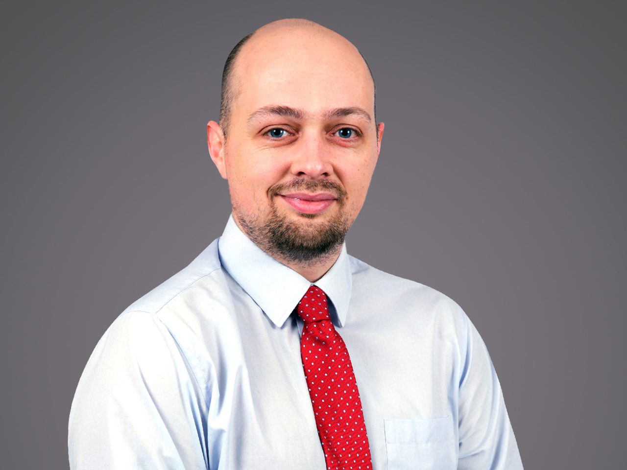 Portrait of a person with a shaved head and light beard, wearing a pale blue shirt and red polka-dot tie, against a grey backdrop.