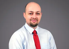 Portrait of a person with a shaved head and light beard, wearing a pale blue shirt and red polka-dot tie, against a grey backdrop.