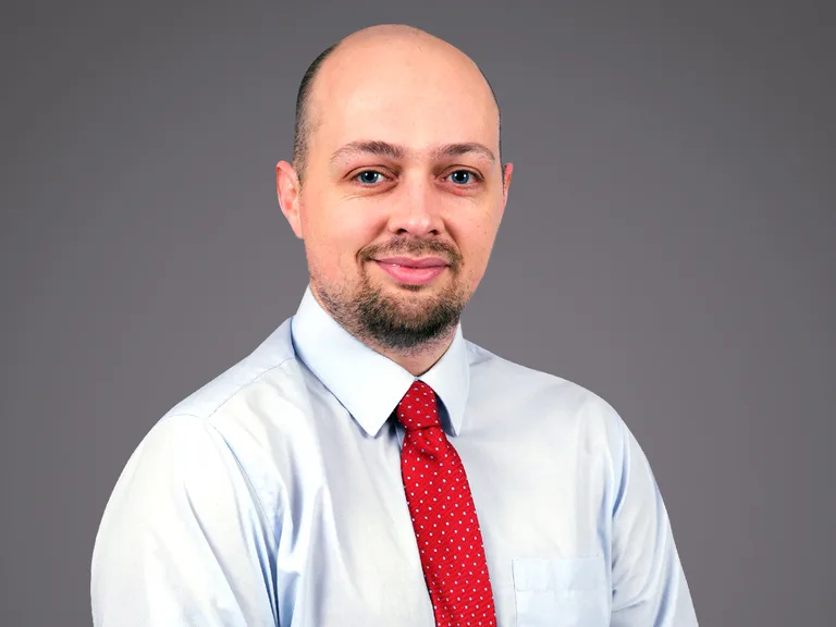 Portrait of a person with a shaved head and light beard, wearing a pale blue shirt and red polka-dot tie, against a grey backdrop.
