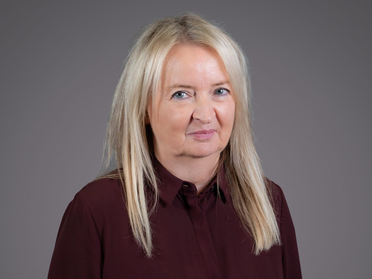 Portrait of a person with long blonde hair wearing a maroon blouse, looking toward the camera with a subtle smile, against a plain gray background.