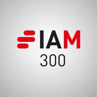 Portrait of a person with short grey hair and glasses, wearing a dark blazer and white shirt, smiling against a plain grey background.