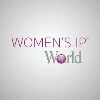 Professional portrait of a person with shoulder-length blonde hair, wearing a black blazer and white blouse, smiling against a grey backdrop.