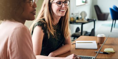 Two women sitting at a table, one with blonde hair and glasses smiling at a laptop, the other with curly hair and glasses, in a bright modern office setting.