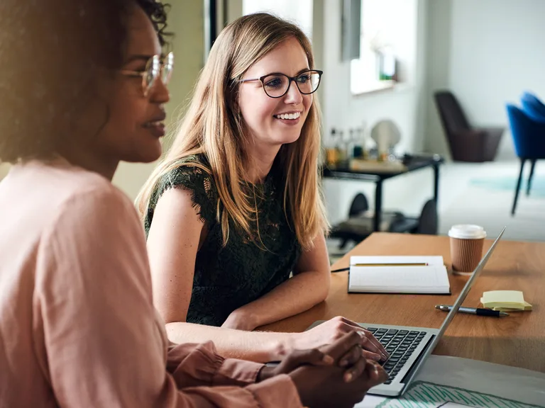 Two women sitting at a table, smiling and working on a laptop in a bright, modern office space with notebooks, coffee, and supplies.
