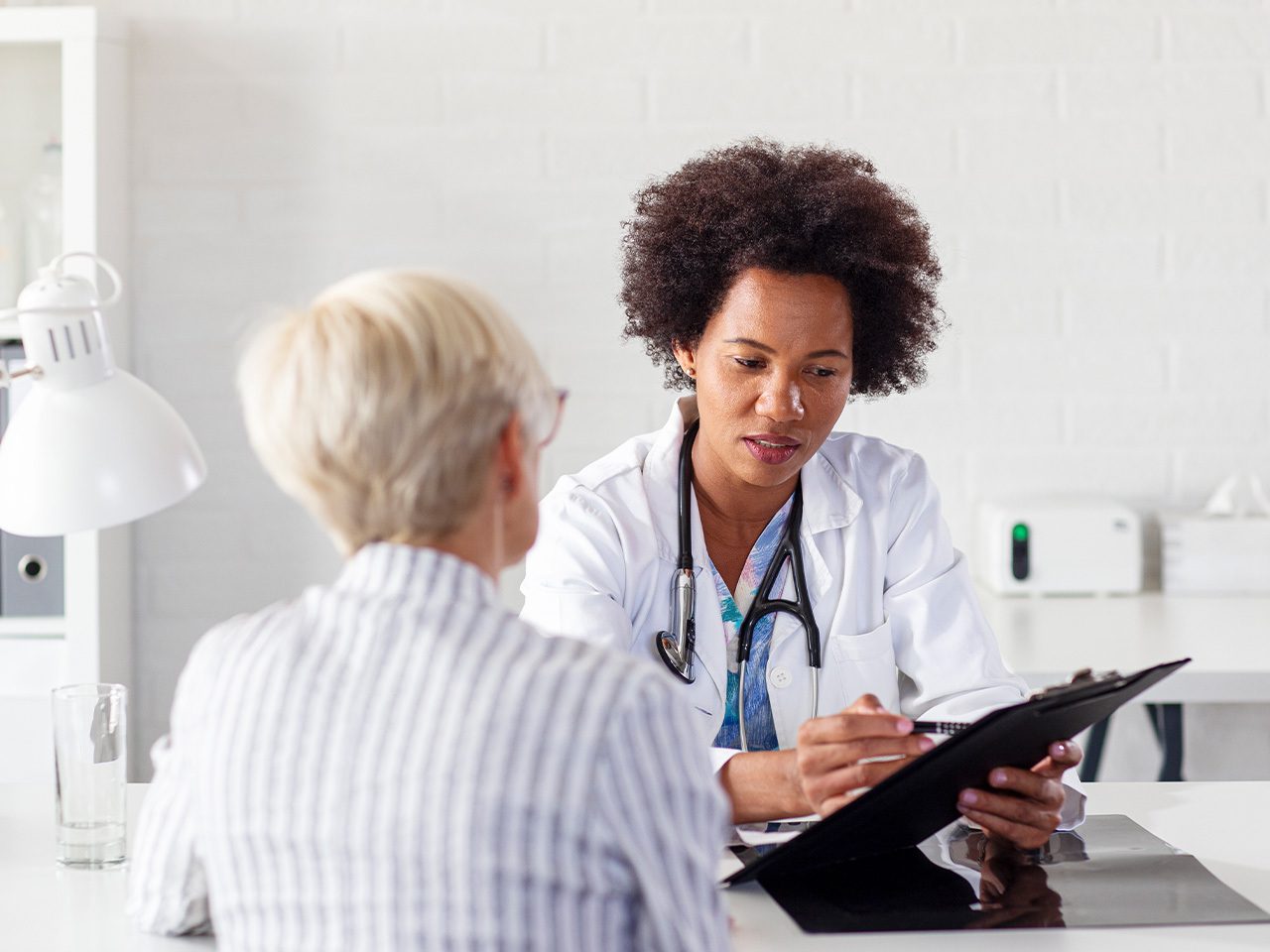 A healthcare professional in a white coat with a stethoscope takes notes on a clipboard while speaking with an older patient at a bright clinic desk.