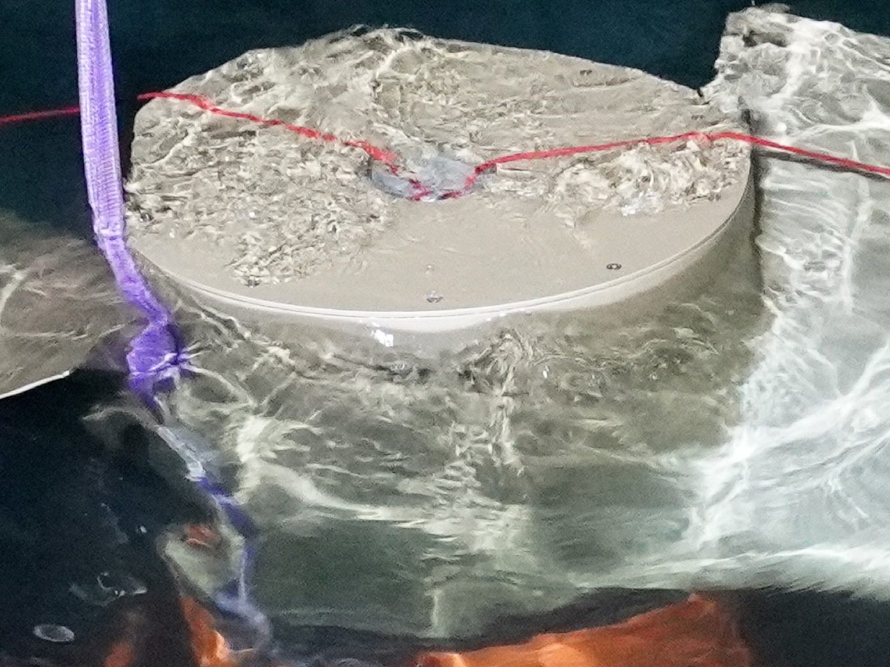 Round grey lid on a crinkled plastic sheet, with a purple strap on the left and a red line stretched across the top.