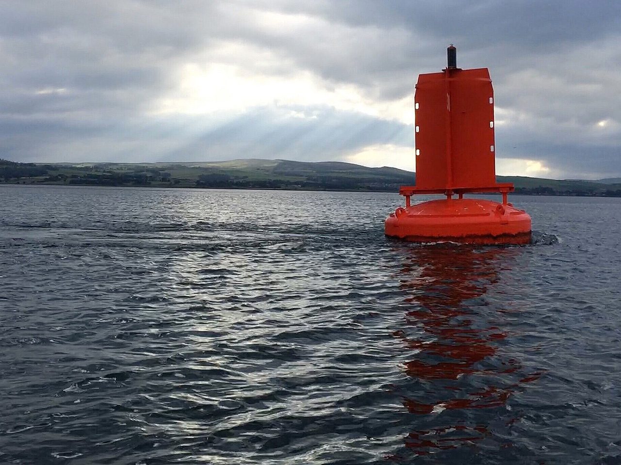Bright orange buoy with a tall vertical beacon sits on a circular base, floating on rippling water with distant hills and a cloudy sky with sun rays.