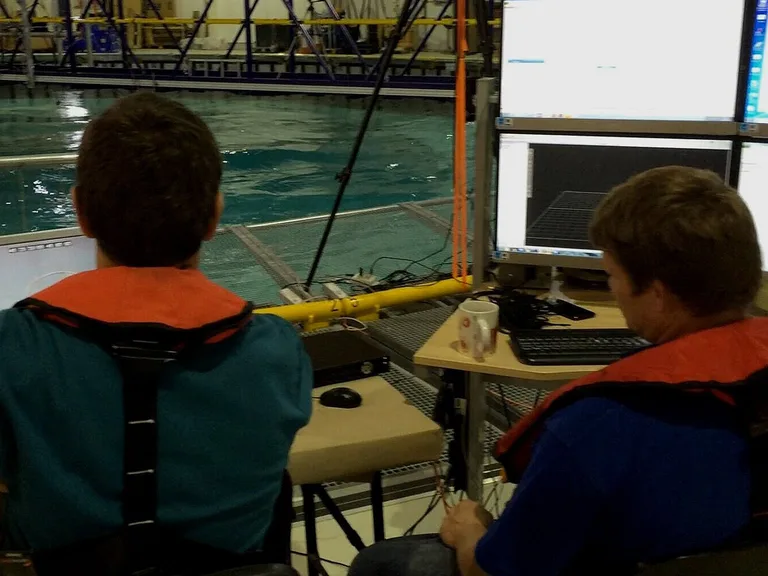 Two people in orange life jackets sit at a control desk with monitors and a keyboard, facing a large pool in a lab or testing facility.