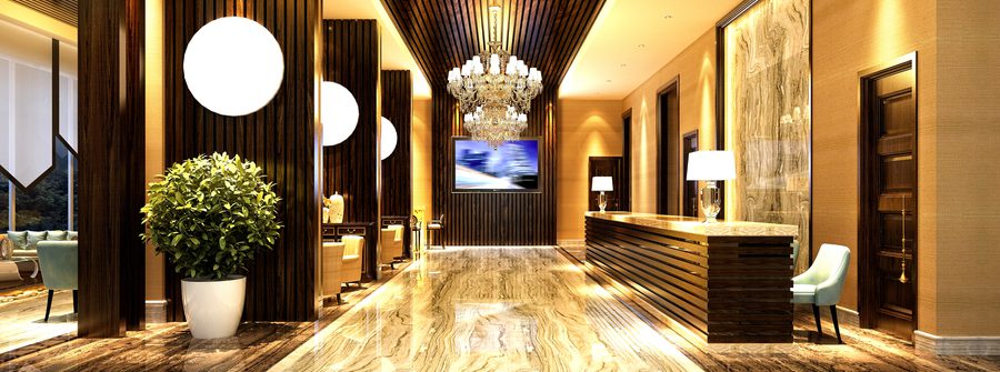 Luxurious hotel lobby with marble floors, dark wood panels, and crystal chandeliers. A reception desk on the right and a seating area on the left, with a potted plant.