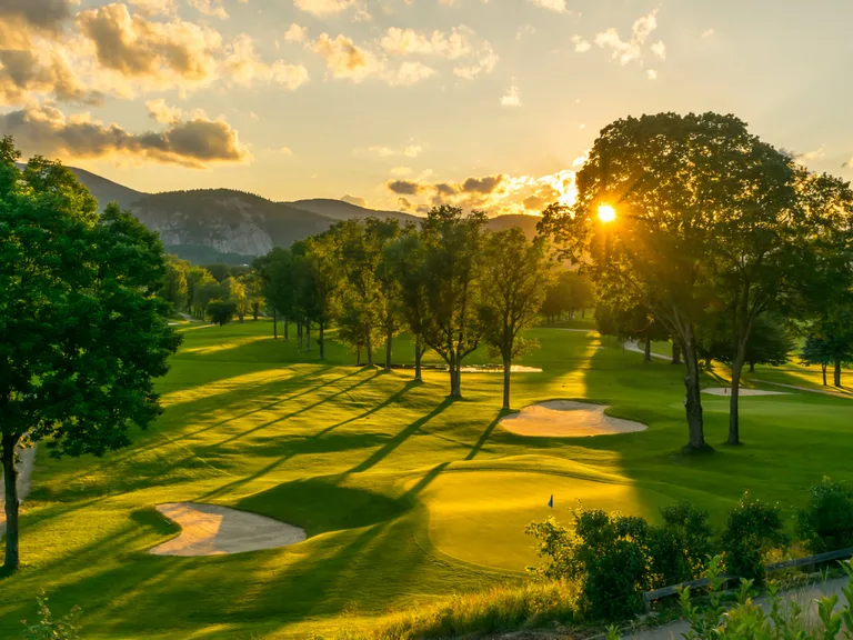 Sunlit golf course at sunset, with long tree shadows, sand bunkers and distant hills under a golden sky.