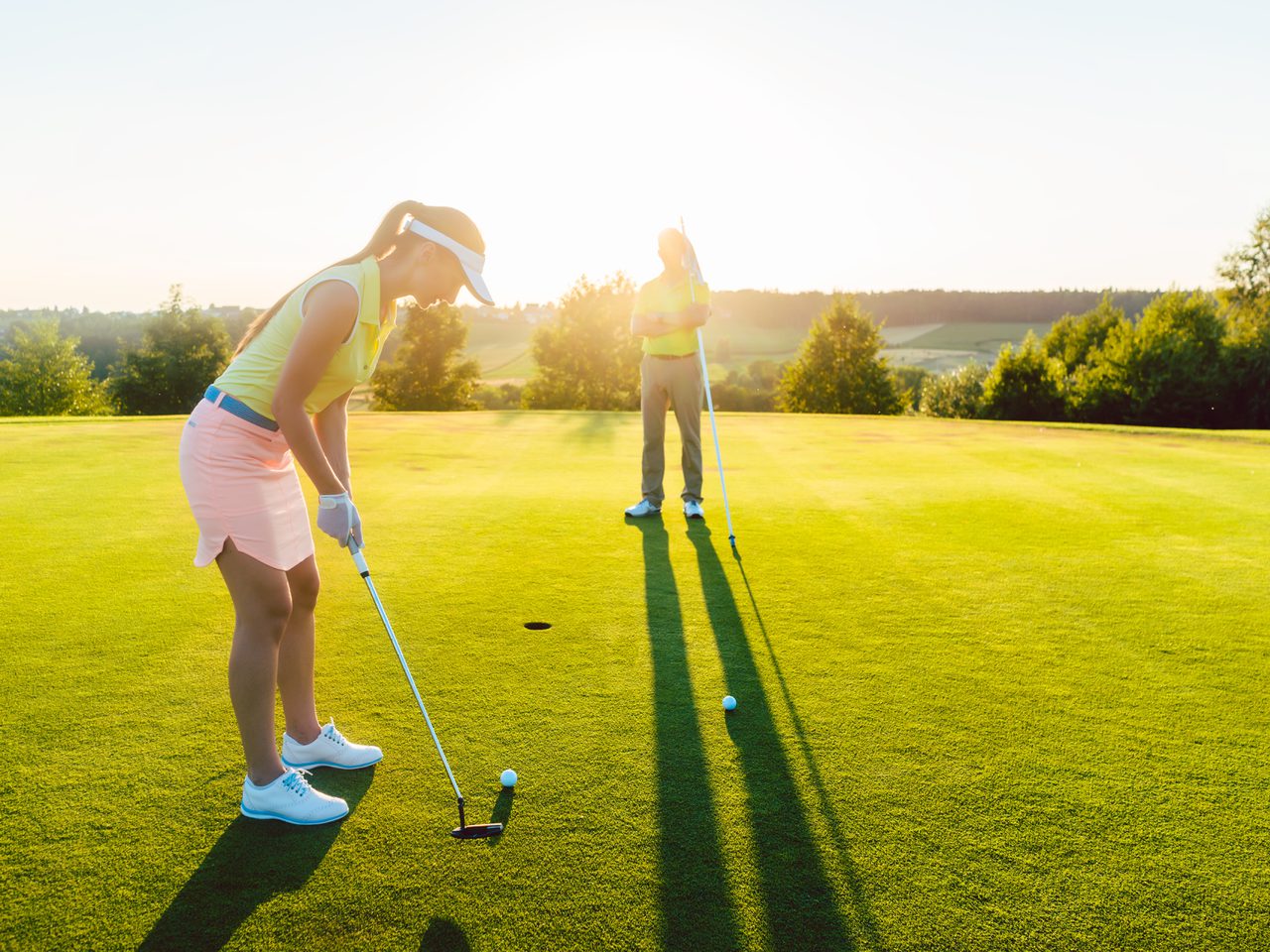 Two golfers on a sunlit golf green at sunset; one bending to putt in a yellow top and pink shorts, the other standing with a club, long shadows cast.