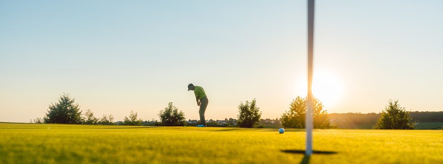 Golfer in a lime-green shirt lines up a putt on a sunlit golf green, with a flagstick casting a long shadow as the sun sets behind trees.